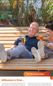 Ein fröhliches Paar sitzt entspannt auf einer Holzbank und genießt ihre Getränke in einer sonnigen Umgebung.
