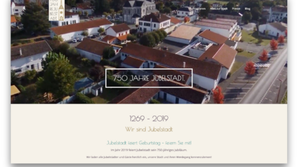 Luftaufnahme einer Stadt mit roten Dächern, umgeben von Bäumen und einer Feier zum 750-jährigen Jubiläum.