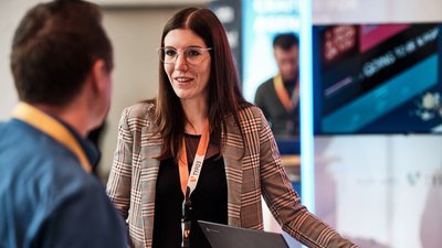 Eine Frau mit Brille und kariertem Blazer spricht mit einem Mann, während sie an einem Tisch mit Laptops stehen.