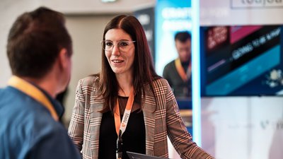 Eine Frau mit Brille und kariertem Blazer spricht mit einem Mann, während sie an einem Tisch mit Laptops stehen.