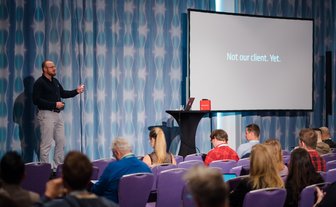 Ein Redner präsentiert auf einer Bühne vor einem Publikum, während eine Folie mit der Aufschrift 'Not our client. Yet.' angezeigt wird.
